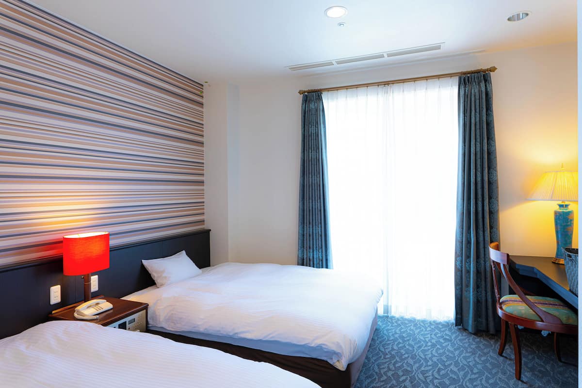 A hotel room with two beds, a desk with a chair, a lamp, and a window with curtains.
