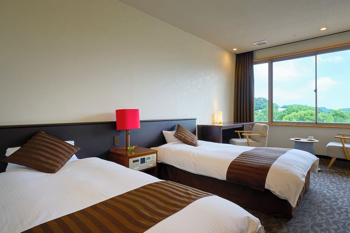 A hotel room with two beds, a desk, a chair, and a large window offering a view.