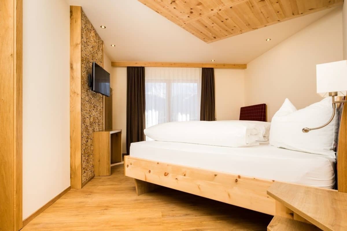 A bedroom with a wooden bed frame, a flat-screen TV mounted on a wooden wall, a bedside table with a lamp, and a window with curtains.