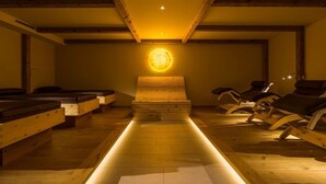 A sauna room with wooden benches and loungers, a central wooden structure, and a warm light source on the ceiling. - Luxapart Ambiente (Ladis)