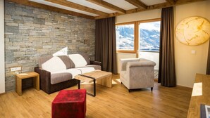 A cozy living room with a stone wall, wooden floor, a sofa, a chair, a red ottoman, and a large window with a mountain view. - Luxapart Ambiente (Ladis)