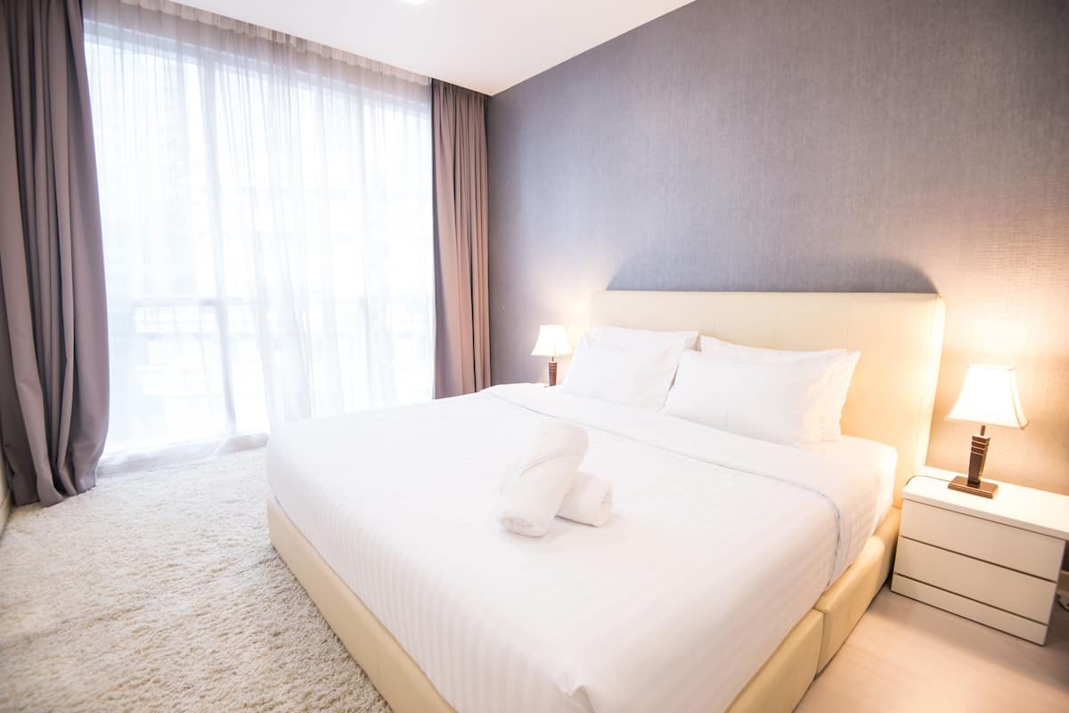 A neatly made bed with white linens and a folded towel, a bedside table with a lamp, and a view of the outdoors through sheer curtains.
