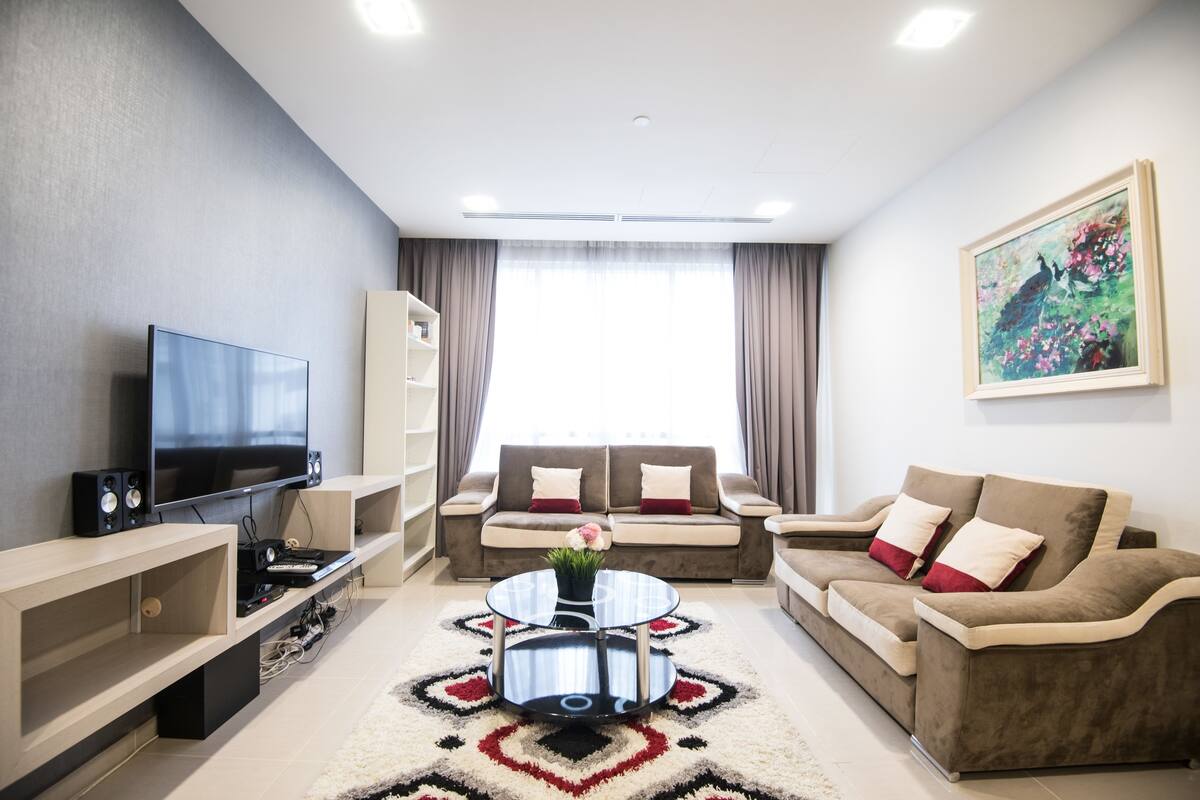 A modern living room with a flat-screen TV, two sofas, a coffee table, and a large window with curtains.