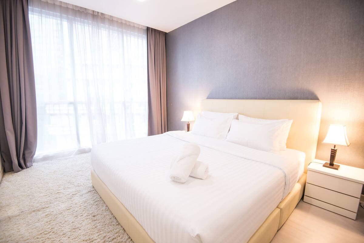 A hotel room with a large bed, two bedside lamps, a nightstand, and a view through sheer curtains.