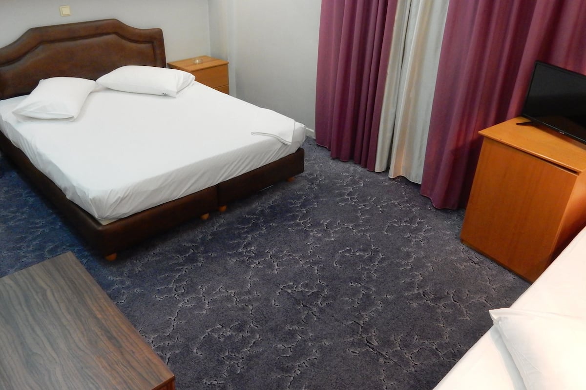 Hotellihuone, jossa on sänky, puinen yöpöytä, televisio puukehikossa ja ikkuna, jossa on verhot.