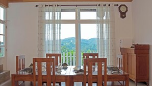 A dining area with a table set for four, wooden chairs, a white tablecloth, and a view of green hills through a large window with bamboo curtains. - Villa Perpetua (Bandarawela)