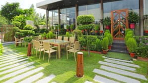 An outdoor seating area with a glass table and chairs, surrounded by potted plants and a well-maintained lawn.