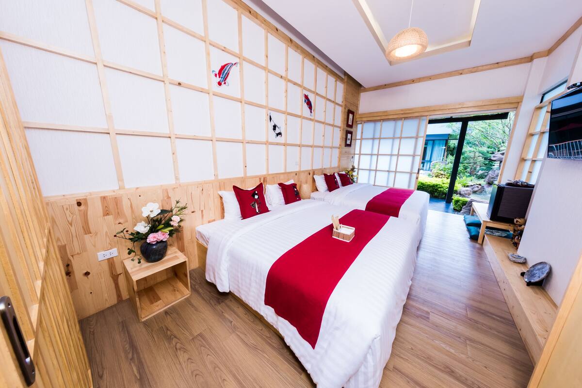 A hotel room with a large bed, wooden paneling on the walls, a small wooden side table with a vase, and a view of the garden through the window.