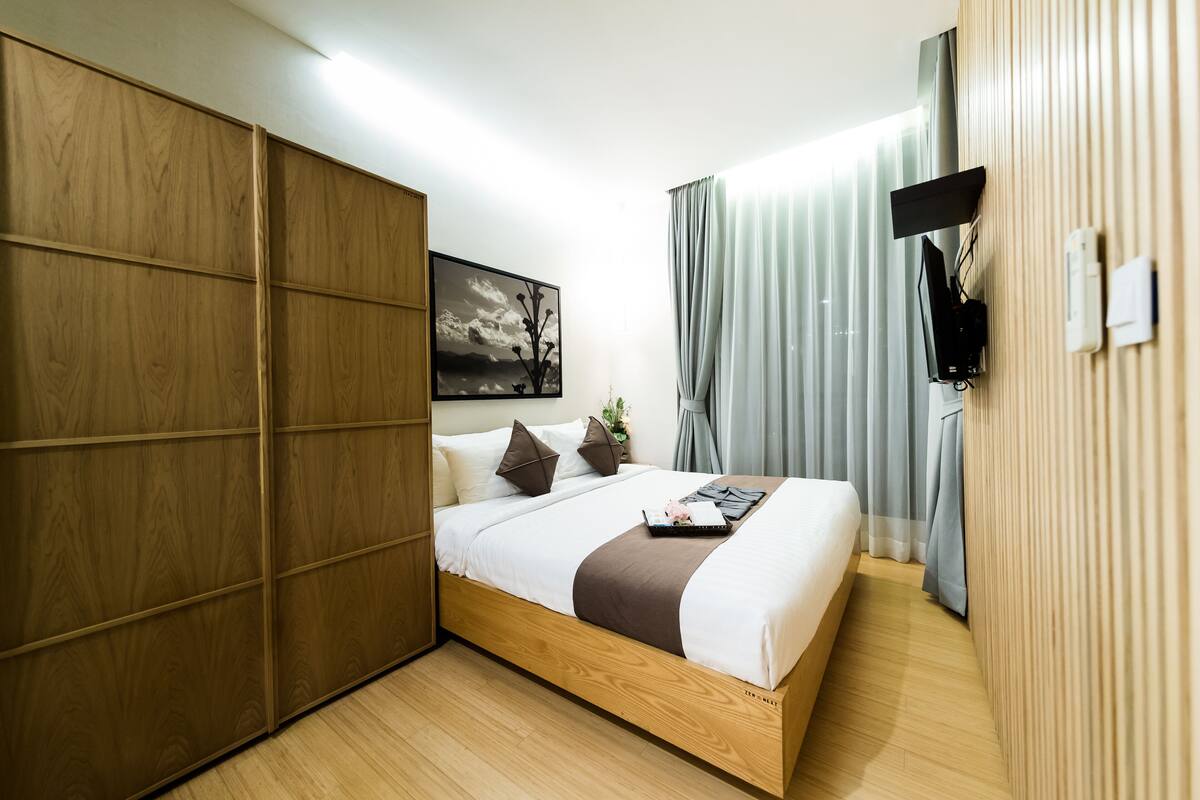 A modern bedroom with a wooden bed frame, a flat-screen TV mounted on the wall, and a large window with curtains.