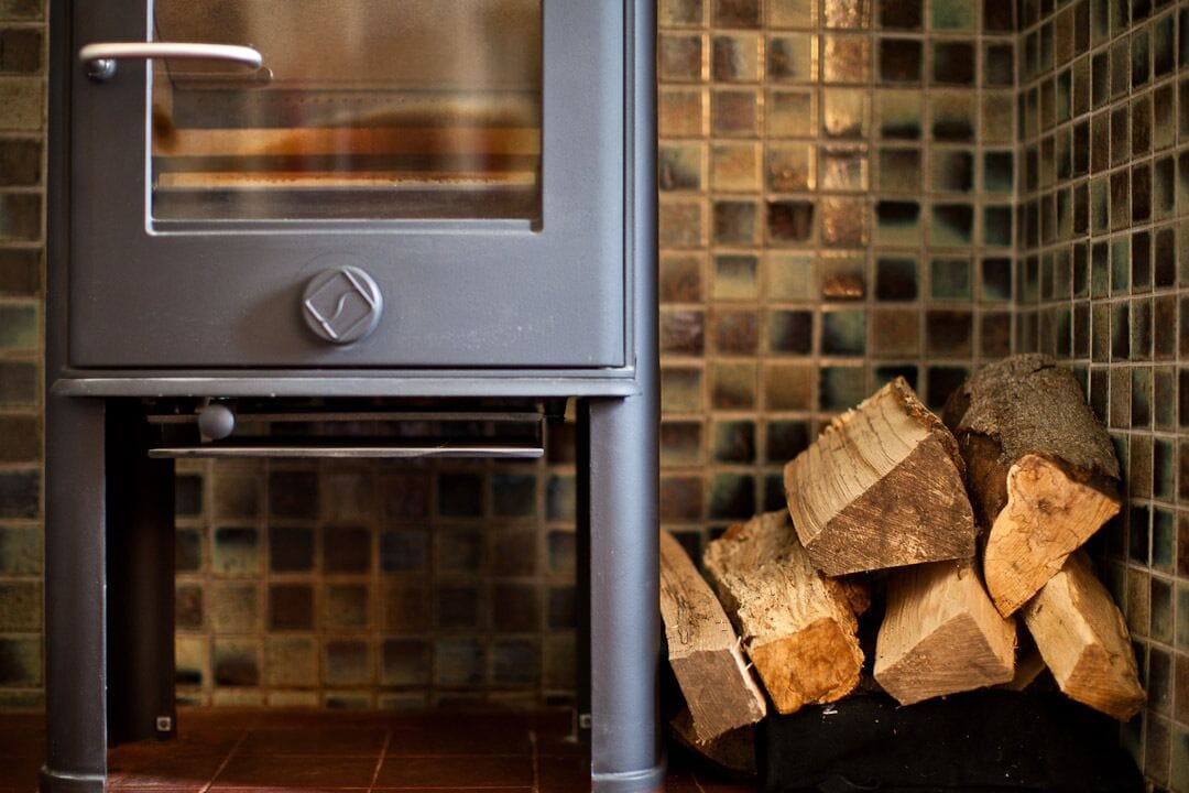 A wood stove with a glass door, a tiled backsplash, and a stack of firewood.
