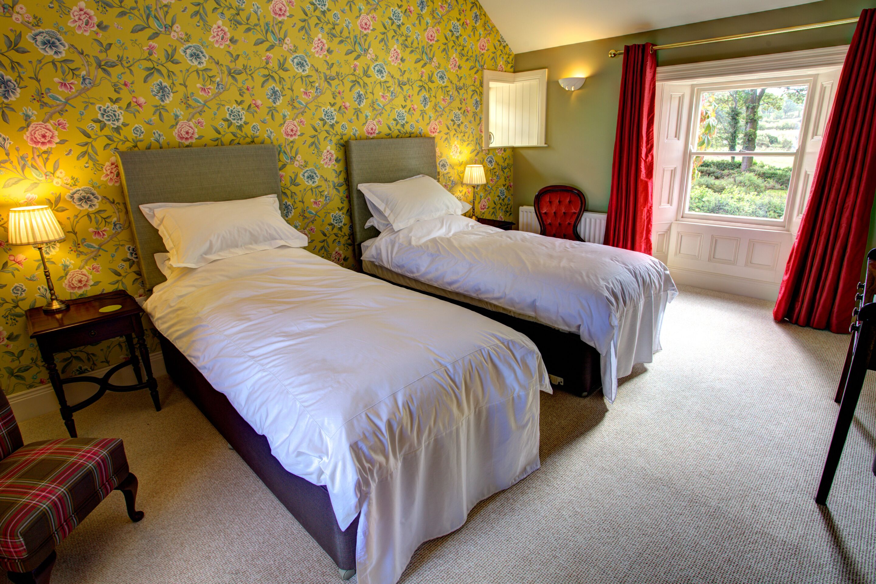 A bedroom with two beds, floral wallpaper, a red chair, and a window with red curtains.