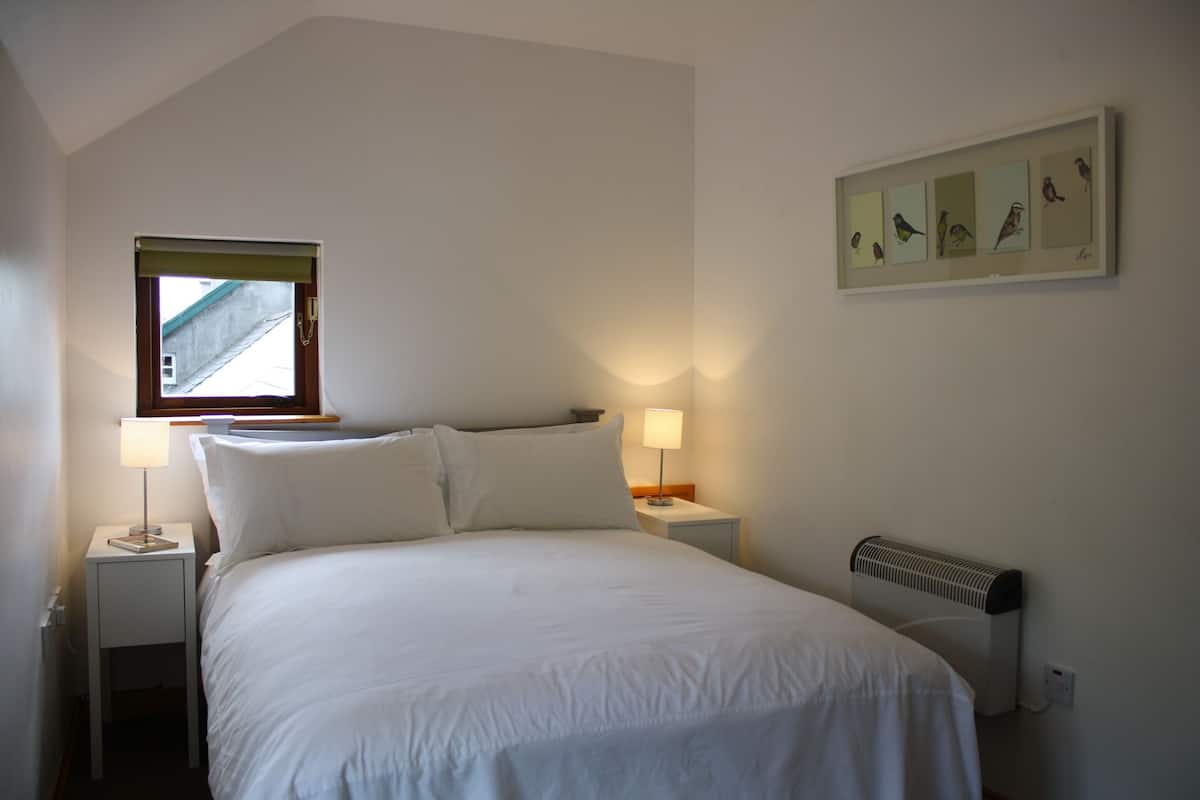A bedroom with a bed, two bedside tables with lamps, a window with a blind, a radiator, and a framed picture of birds on the wall.
