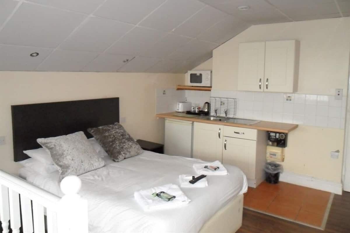A compact bedroom with a bed, bedside table, and a kitchenette area including a microwave and sink.