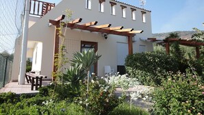 Une maison à deux étages avec une pergola, un patio doté d’un banc et un jardin où poussent différentes plantes et fleurs.
