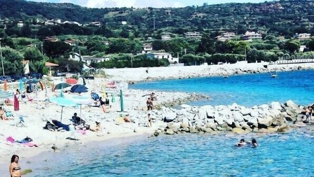 Une plage avec une eau cristalline, des gens qui se baignent et prennent des bains de soleil, et des maisons en arrière-plan.