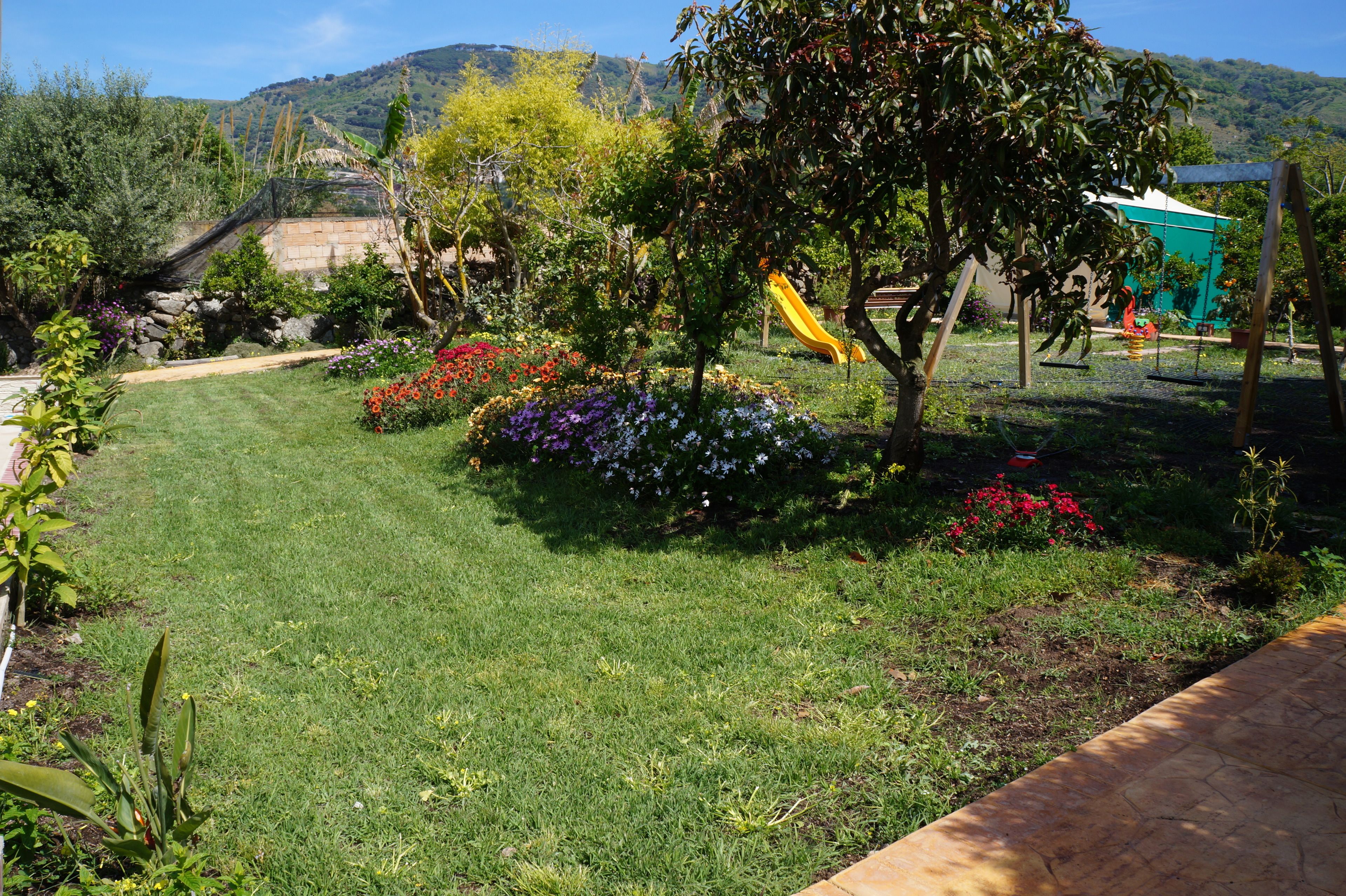 Un jardí ben cuidat amb gespa, llits de flors de colors i una zona de jocs que inclou un tobogan.