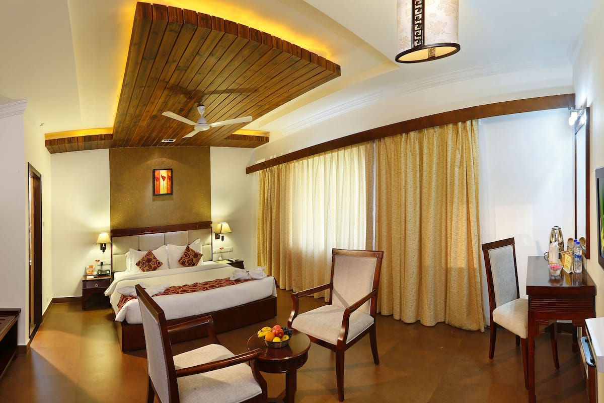 A hotel room with a large bed, a wooden ceiling, a ceiling fan, a desk with a chair, and a window with curtains.