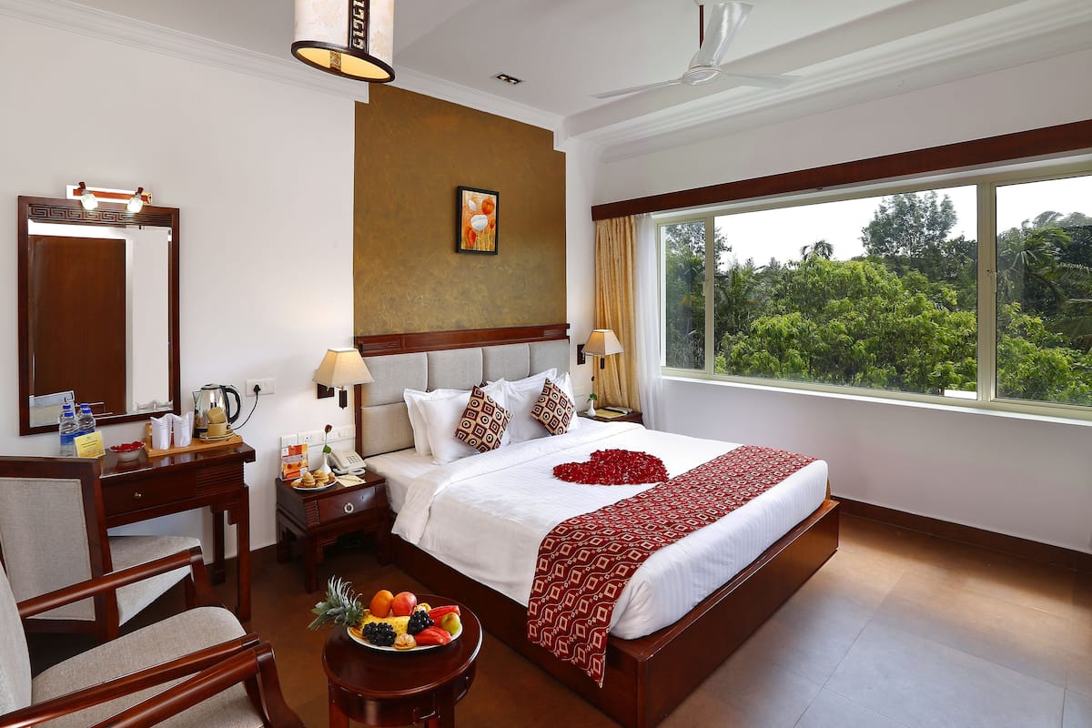 A hotel room with a large bed, a desk with a mirror, a chair, a bedside table with a lamp, and a window with a view of greenery.