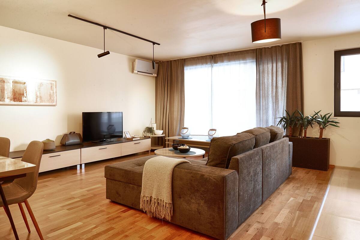 A modern living room with a sofa, TV, dining table, and a large window with curtains.