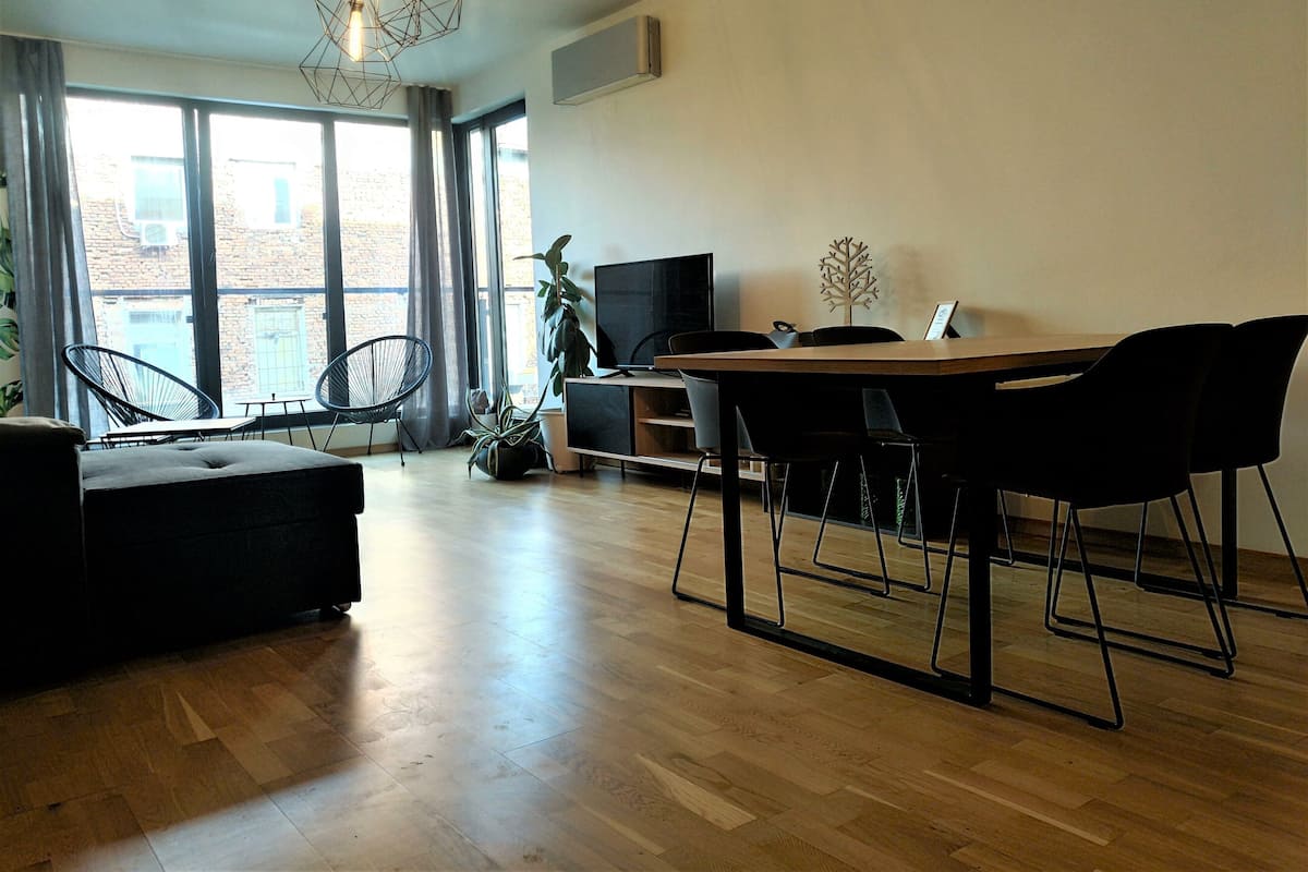 A modern living room with a wooden floor, a dining table with chairs, a sofa, a TV, and a plant.