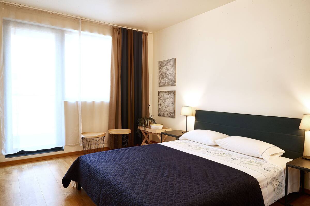 A hotel room with a bed, a bedside table, a lamp, a small table with a bowl, and a chair.