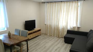Una sala de estar con una mesa de comedor de madera, un televisor sobre un mueble de madera, un sofá gris y una ventana con cortinas.