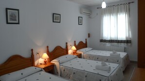 Une chambre d’hôtel avec trois lits simples, chacun doté d’une tête de lit en bois, d’une lampe de chevet et d’une couvre-lit blanche ornée d’un motif de feuilles. On y trouve une fenêtre avec des rideaux blancs, un climatiseur fixé au mur et des tableaux encadrés accrochés au mur.