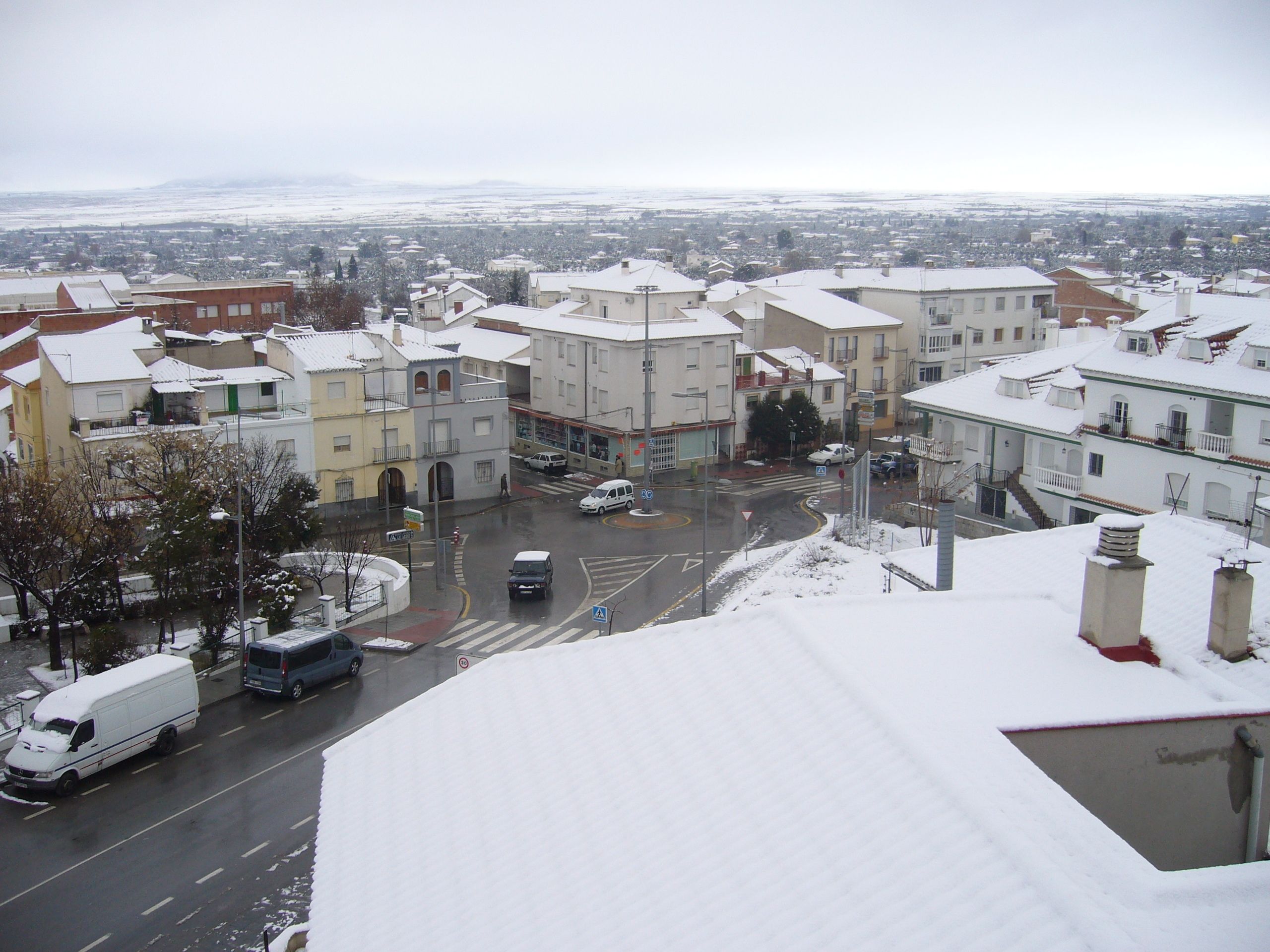 A snow-covered urban street with buildings, a pedestrian crossing, and vehicles.