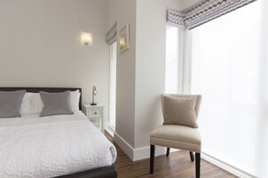 Un dormitorio con cama, mesita de noche, lámpara, silla y ventana con cortinas.