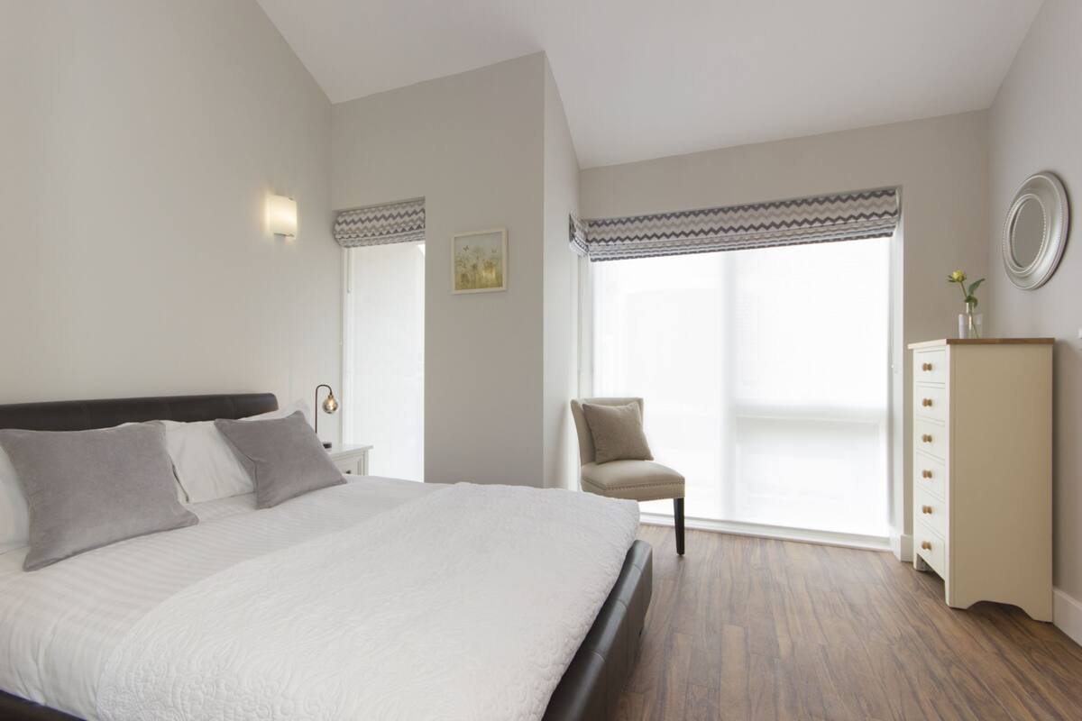 Une chambre à coucher avec un grand lit, une chaise, une commode et une fenêtre avec des rideaux.