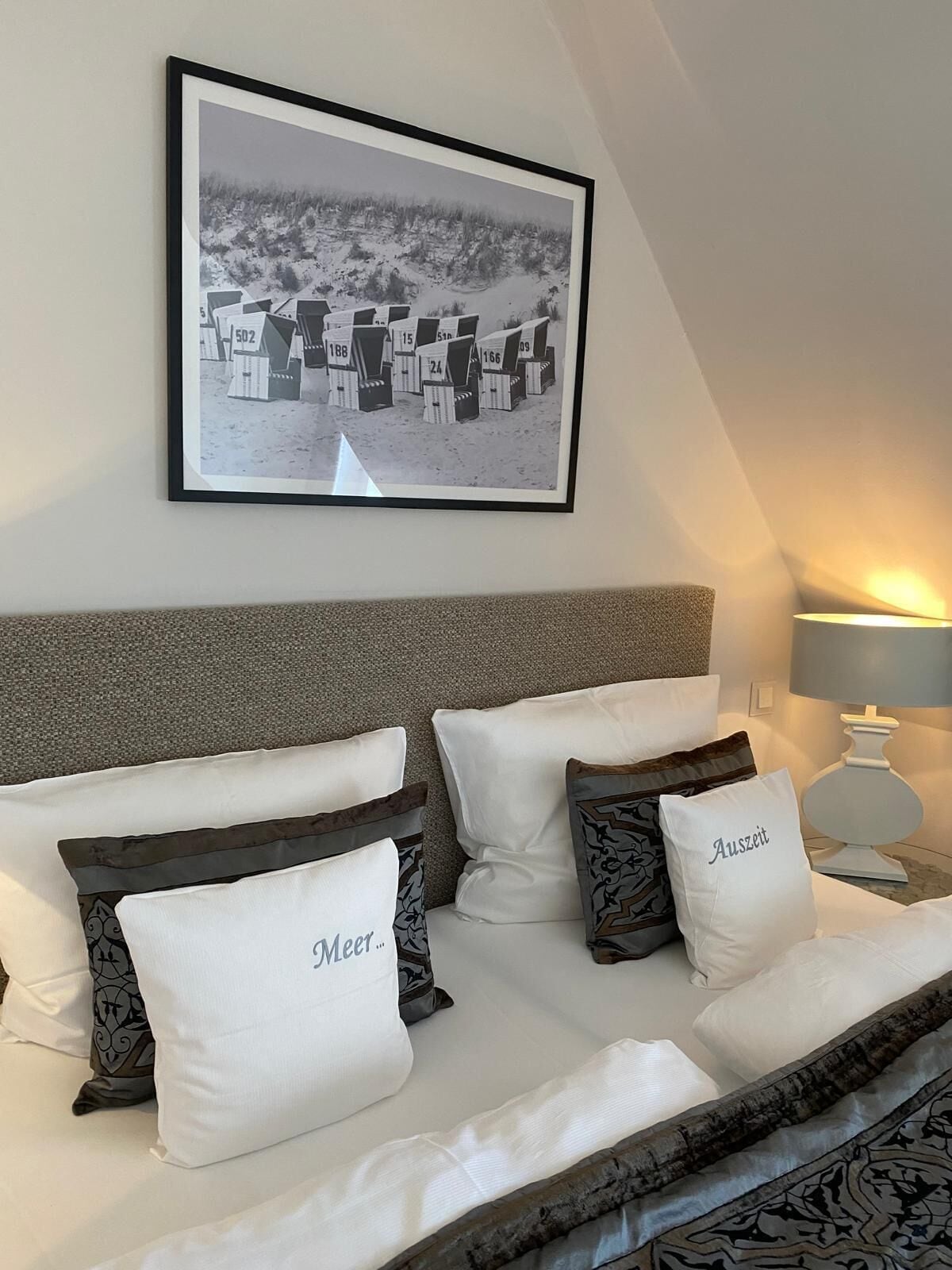 A neatly made bed with white and patterned pillows, a framed photograph above it, and a bedside lamp.
