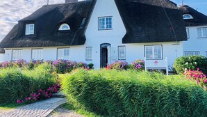 A traditional white building with a black thatched roof, surrounded by well-maintained hedges and flowering plants. - Strandvogtei Sylt (Sylt)