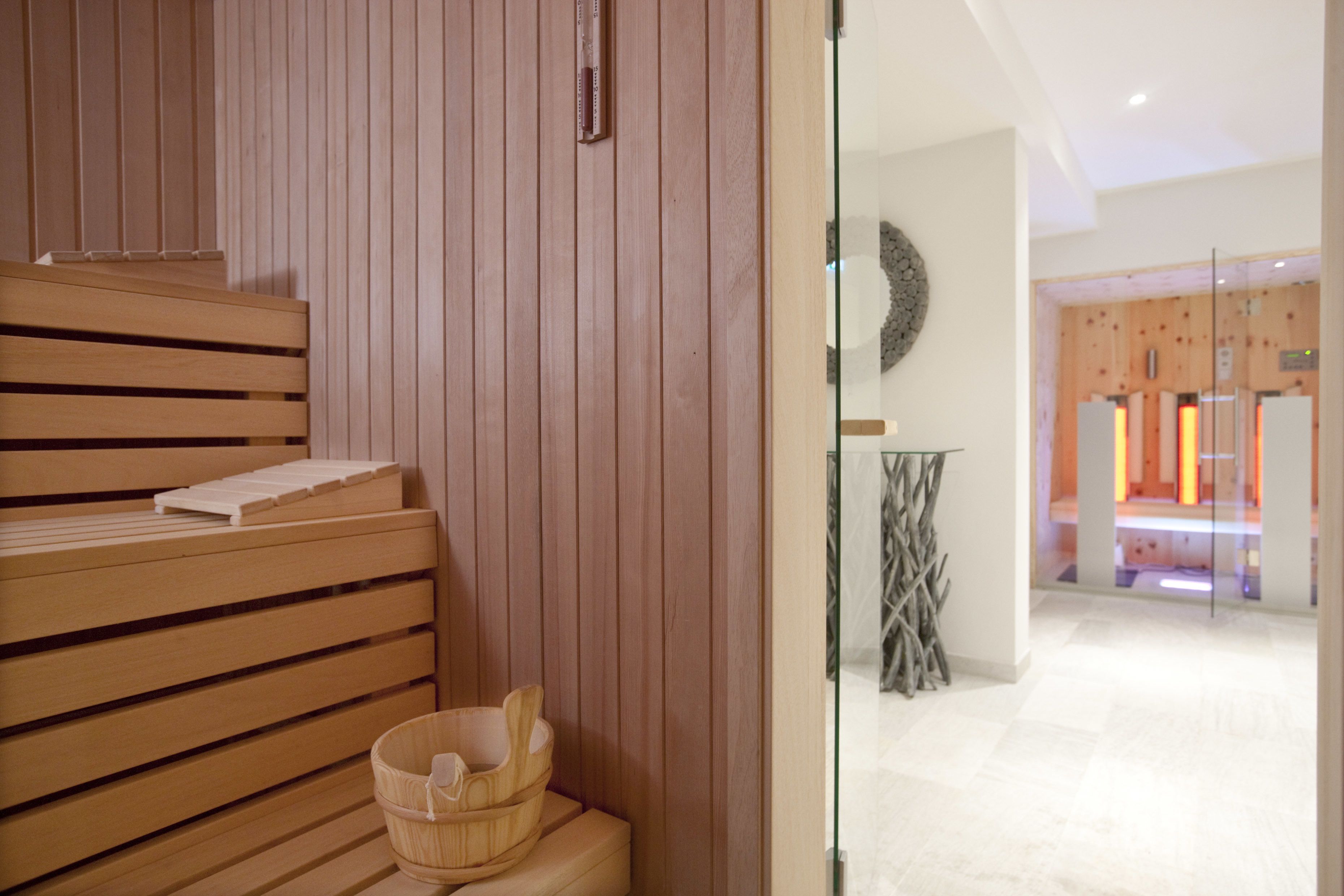A wooden sauna bench with a wooden bucket, a glass partition, and a modern interior with a tiled floor and wooden paneling.
