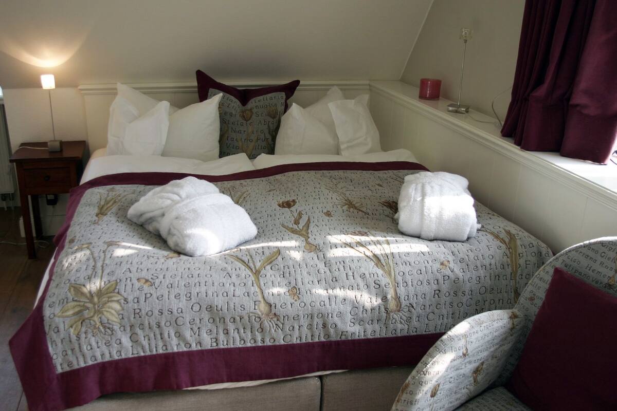 A neatly made bed with a floral-patterned bedspread and two towels, a bedside table with a lamp, and a window with curtains.