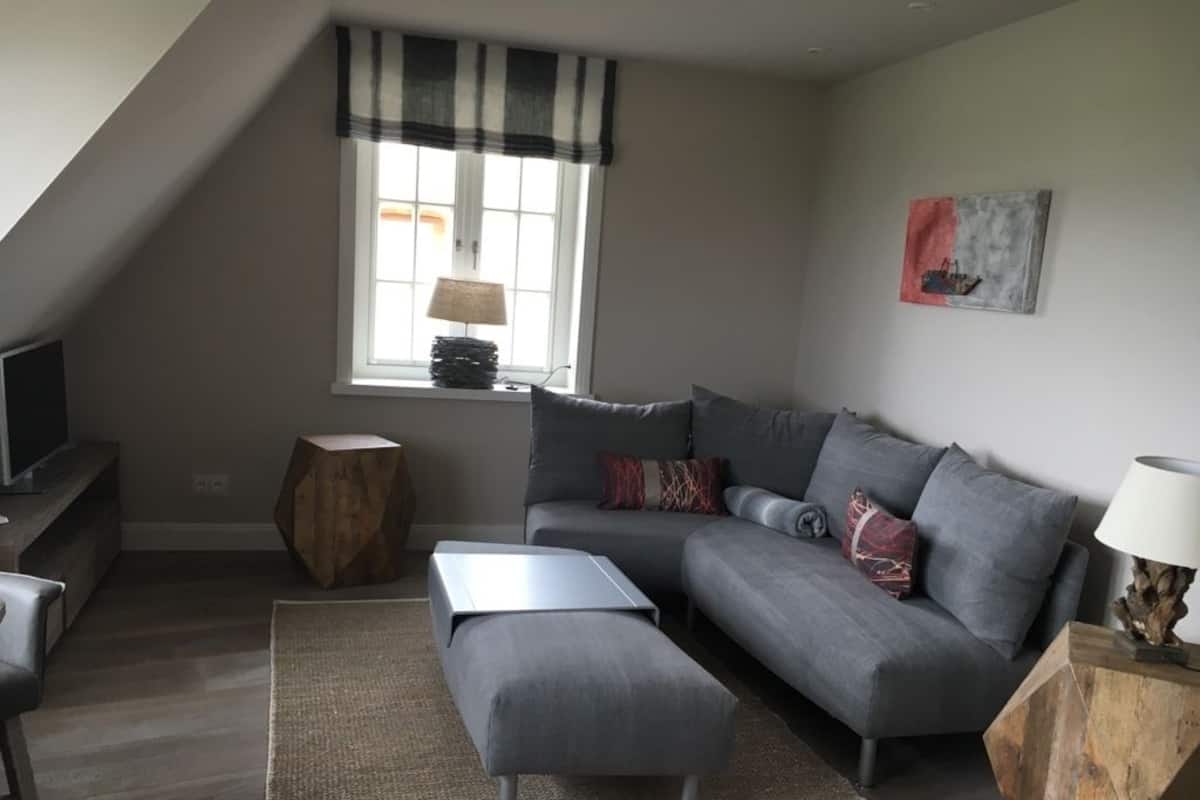 A modern living room with a grey sofa, a wooden side table, a small wooden stool, and a TV on a stand.