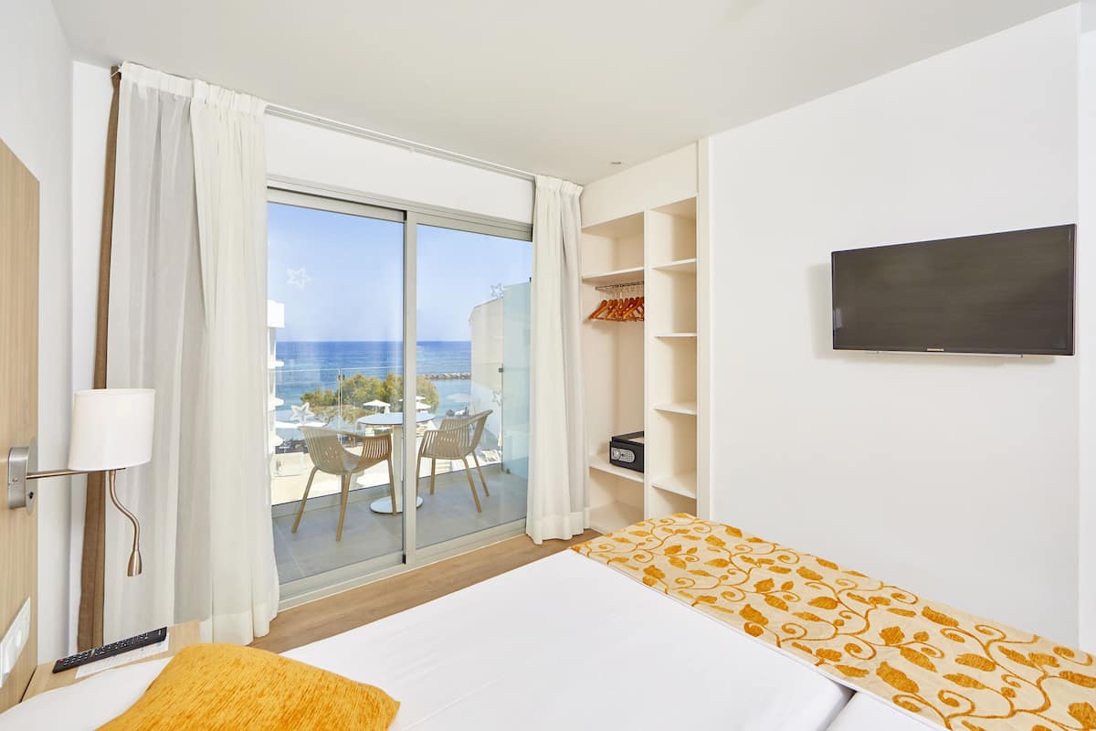 A hotel room with a bed, a TV, a wardrobe, and a balcony with a view of the sea.