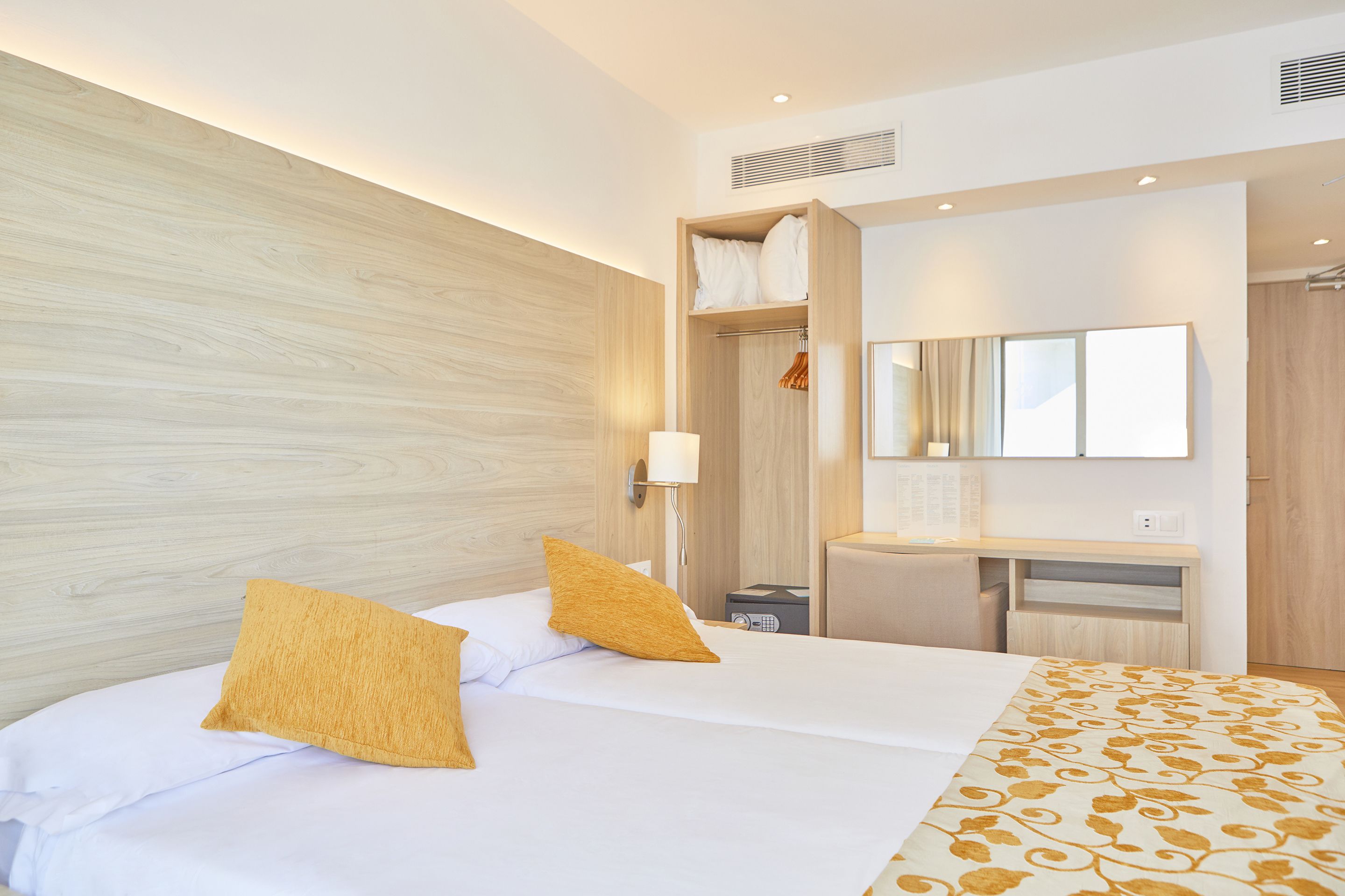 A modern hotel room with a wooden headboard, a bed with white linens and two mustard-colored pillows, a bedside table with a lamp, a built-in desk with a mirror, and a wardrobe.