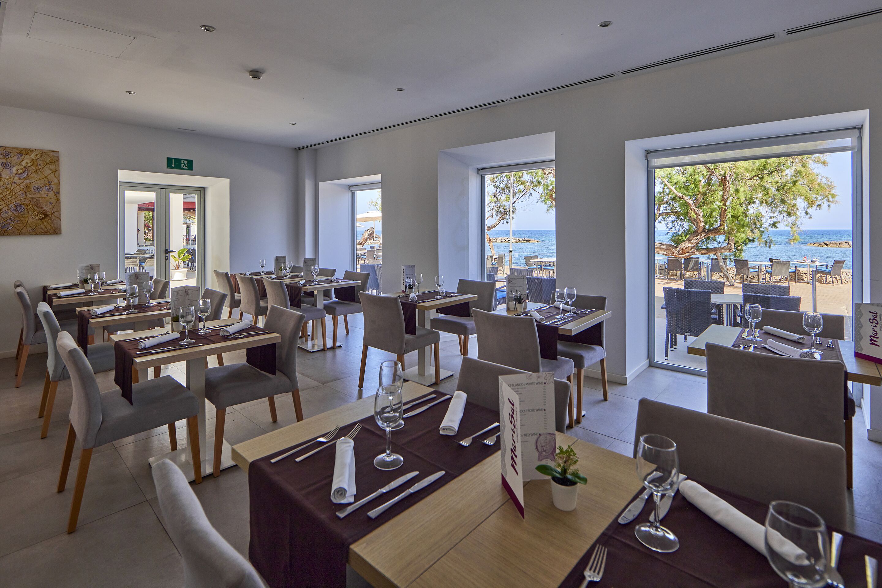 Un restaurante con mesas preparadas para comer, amplias ventanas que ofrecen vistas al mar y un letrero que indica la salida.