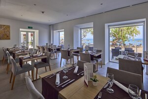 Un restaurante con mesas preparadas para comer, amplias ventanas que ofrecen vistas al mar y un letrero que indica la salida.