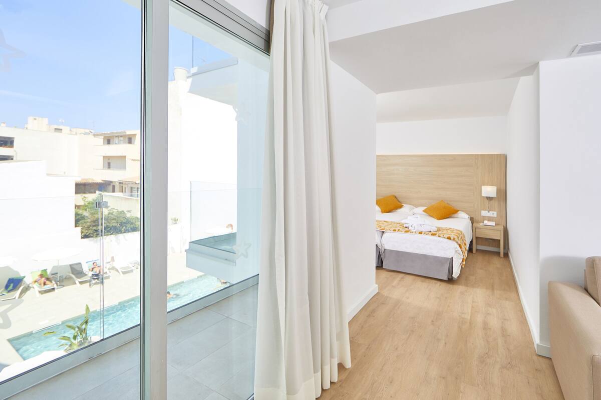 A hotel room with a large window, a bed with yellow pillows, a wooden headboard, and a bedside table with a lamp.