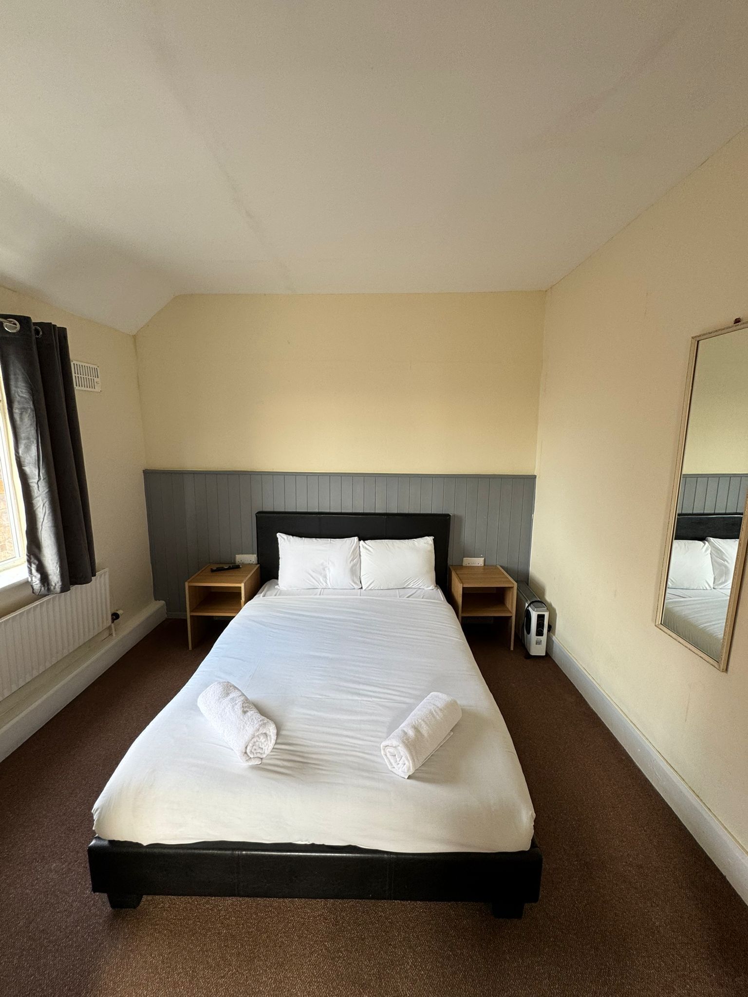 A hotel room with a large bed, two bedside tables, a heater, a mirror, and a window with curtains.