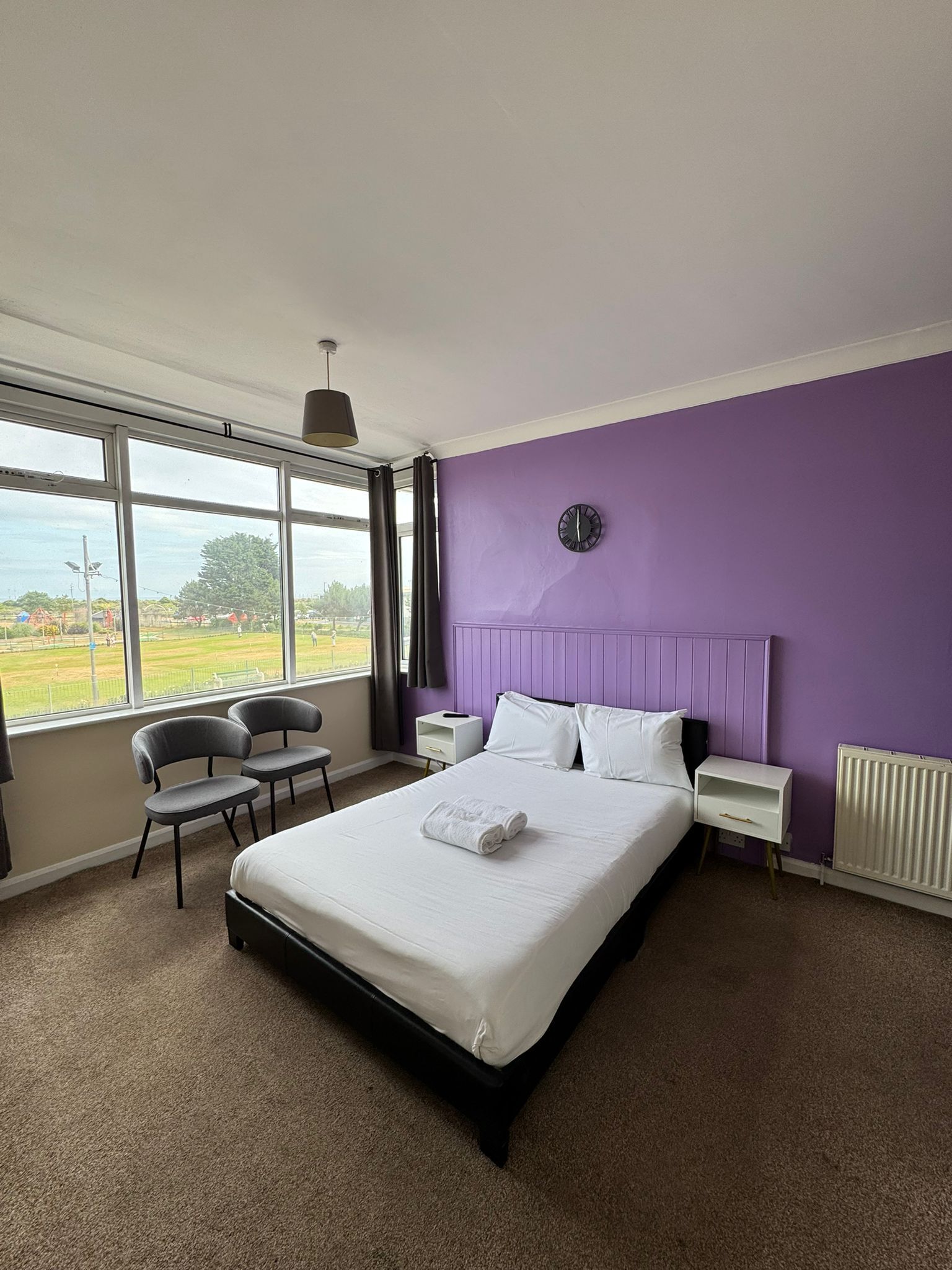 A bedroom with a large bed, a chair, a nightstand, a clock, and a view of a field through the window.