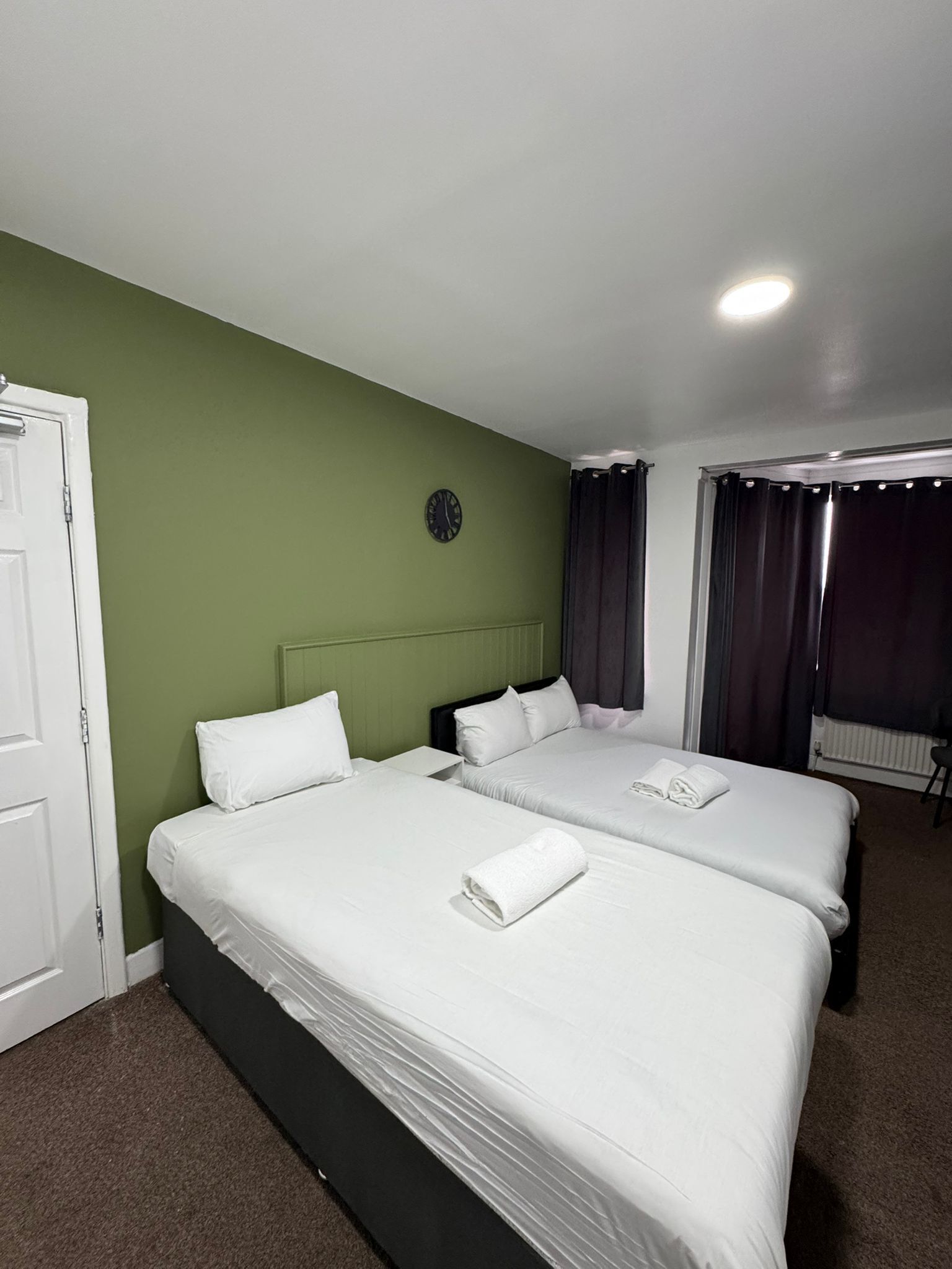 A hotel room with two beds, green walls, a clock, and a window with curtains.