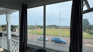A room with two chairs and a table, large windows with curtains, and a view of a road and greenery outside. - Sea Side Hotel (Skegness)