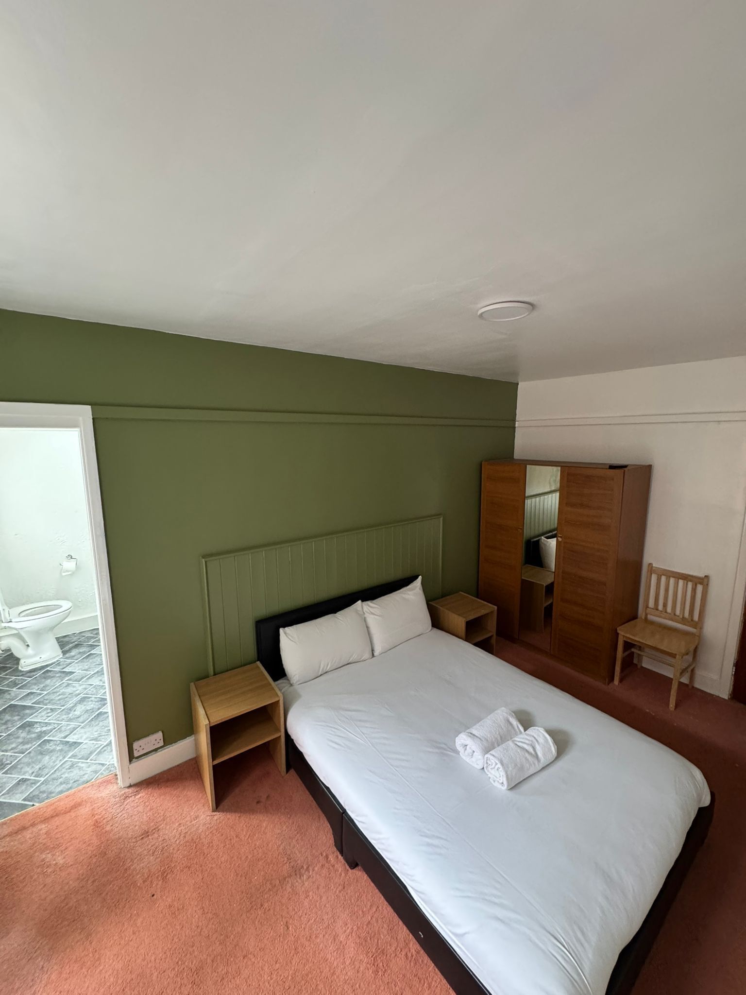 A hotel room with a bed, a wooden nightstand, a wooden chair, a wardrobe, and a bathroom visible through a glass door.