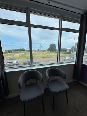 A room with three windows, two chairs, and a view of a park and parking area. - Sea Side Hotel (Skegness)