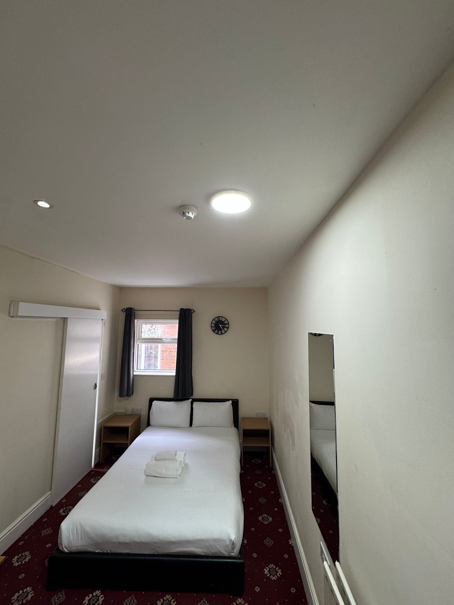 Une chambre d’hôtel avec un lit simple, une table de chevet, une petite table de nuit en bois, une fenêtre avec un rideau, une horloge murale et un luminaire au plafond.
