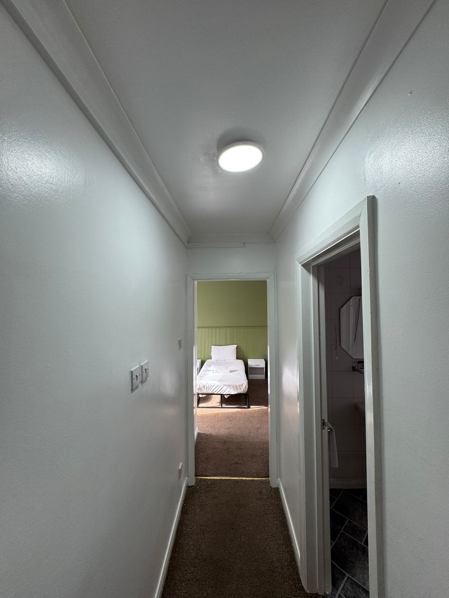 Un pasillo estrecho que conduce a un dormitorio con cama, mesitas de noche y una puerta a otra habitación.