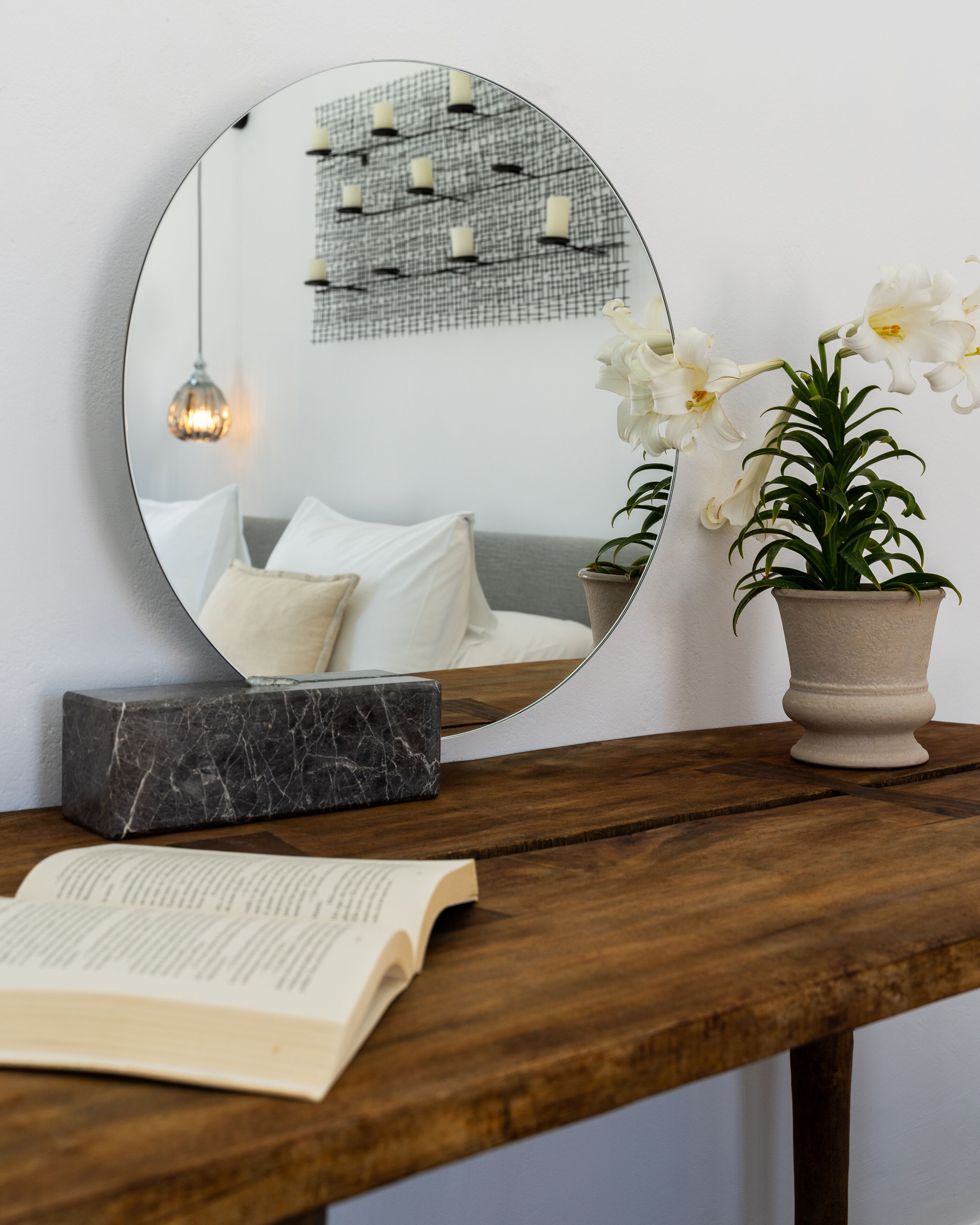 A round mirror on a wooden table, reflecting a room with a bed, pillows, a hanging light, and a potted plant.