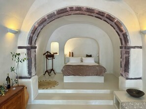 A bedroom with a bed, a wooden dresser, a small table, and a window. - Elysian Santorini (Santorini)